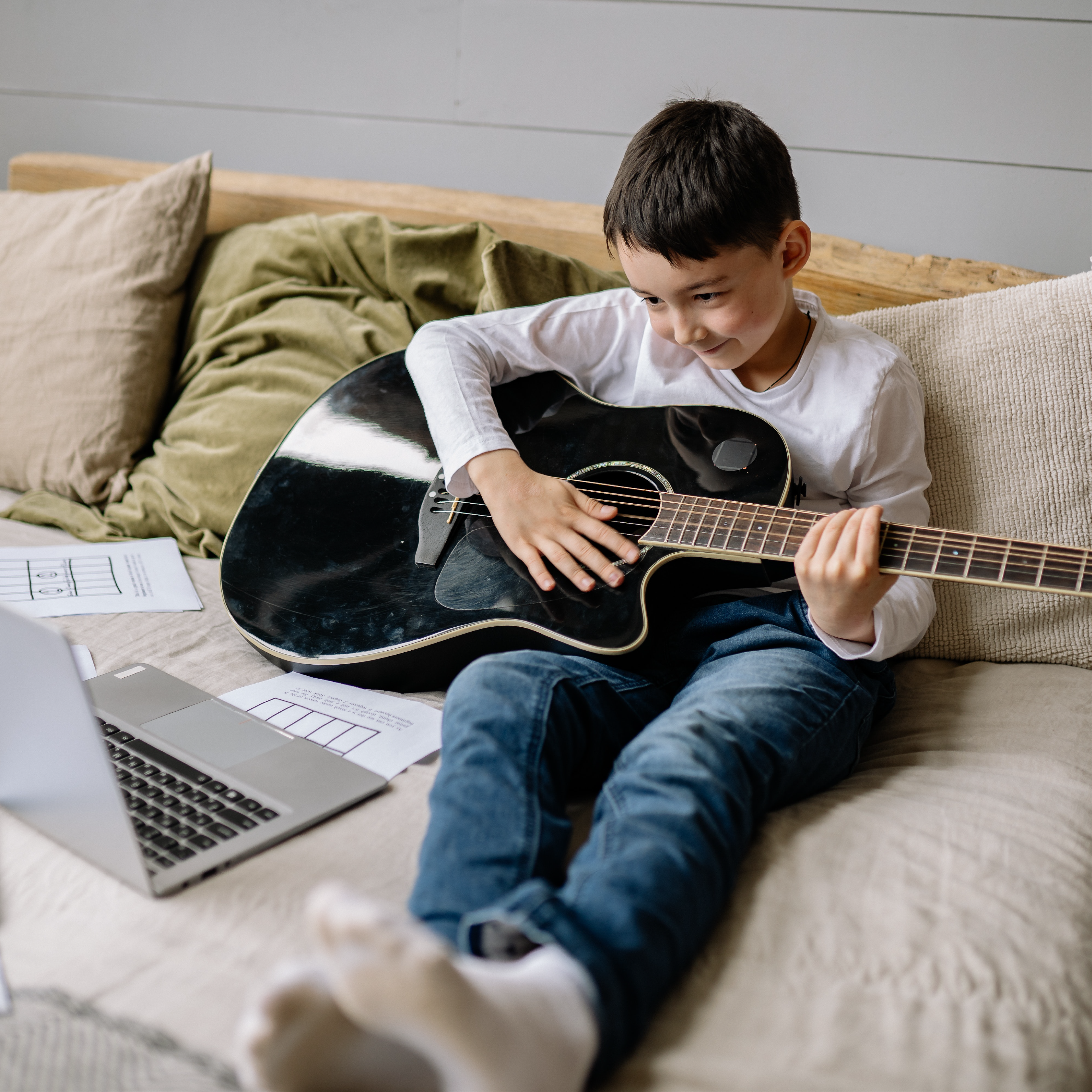 Boy Sitting on Bed with Acoustic Guitar Smiling at a Laptop by Yan Krukau